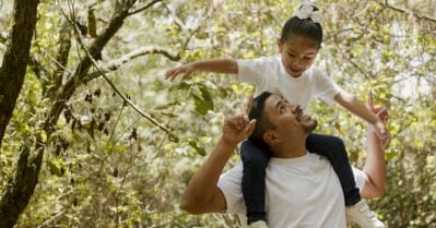 A young Latino father smiles up at his toddler-age daughter, who sits atop his shoulders.