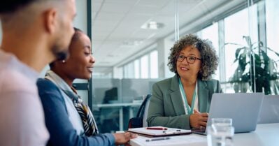 In an office setting, a middle-aged woman of color sits at a table with a laptop in front of her. Seated next to her — also smiling — is a young Black woman, along with a young man.