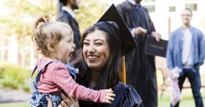 A young adult mother, in her cap and gown, smiles at her baby girl. The sit on a lawn, with other graduates in the background.