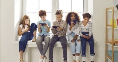 A group of children of different races and ethnicities sit in a row on a windowsill. Their faces show no emotion as they stare at their mobile phones.