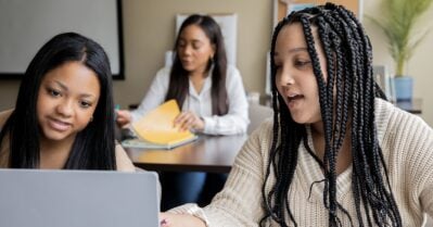 Two young black women are in class, looking at a laptop and engaged in discussion.