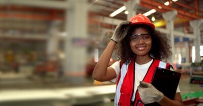 A young black women is working in a manufacturing plant. She is wearing a construction vest, hard hat and clipboard and smiling at the camera.