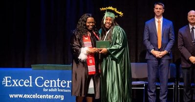 An Excel Center graduate stands on stage, smiling in a cap and gown, as she is being handed her high school diploma.