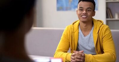 A young man of color smiles while sitting at a desk, his hands clasped in front of him, in a bright yellow hooded sweatshirt.