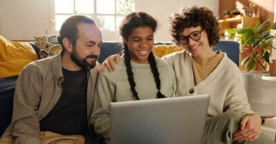 A Black adolescent girl smiles, while sitting between two white foster parents. The three of them are looking at a laptop held in the girl’s lap.