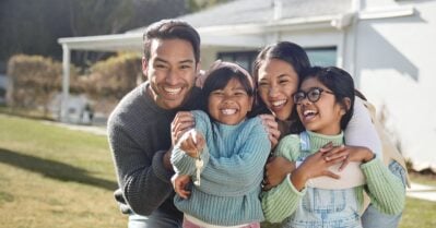 A family of color smiles and squishes together for a family photo outside. All four members are on the lawn outside a white house.