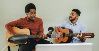 Two young Black men sit together as they practice playing guitar.