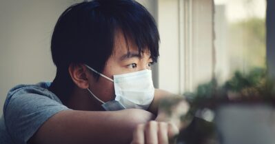 A young asian man looks out the windoww, his chin rests on his folded arms on the ledge; he's wearing a mask to prevent the spread of COVID-19.