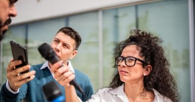 A male and a female journalist point microphones and tape recorders toward a male subject.