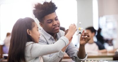 A Black teenage boy tutors a young Latin girl in science. They hold a model of a molecule and discuss it in a classroom of other students.