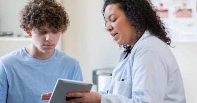 A female doctor in a white medical coat holds up an iPad and shows it to a young male patient in her care.