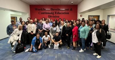 A group of diverse people fill a room, standing in front of a wall displaying "Workforce Development and Continuing Education Division"