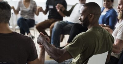 A diverse group of adults sit in a circle and hold hands in unity.