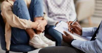 A counseling session in progress. In the foreground, a professional with dark skin is holding a clipboard and pen, taking notes. Two individuals sit across from the therapist — one has knees drawn up to the chest and wears jeans and sneakers, suggesting a potentially distressed or vulnerable posture. The other wears a light sweater vest and jeans. The setting appears calm and professional.