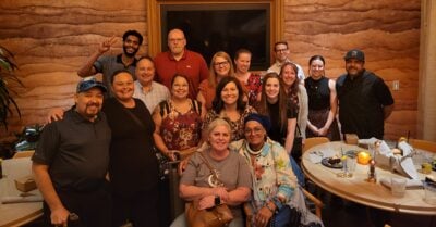 A group of sixteen people gather closely together in a warmly lit restaurant, smiling for a group photo. Some stand while others sit around a table with plates, glasses and candles, creating a relaxed and cheerful atmosphere.