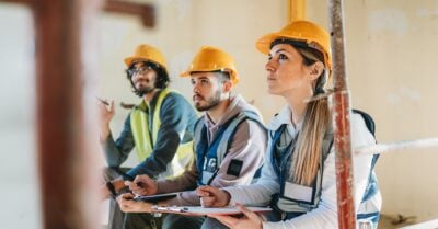 Three young adults wearing safety vests and yellow hard hats sit indoors at a construction site, attentively taking notes during a training session.