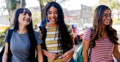Three young ladies walk outdoors, all wearing backpacks and smiling.