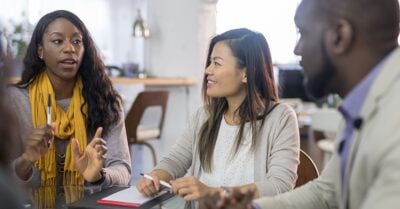 Three colleagues sit around a table in a meeting, with one woman speaking and gesturing while the others listen and take notes.