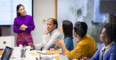 A group of professionals seated around a conference table participate in a staff meeting while a colleague presents at the front of the room.