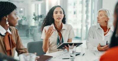 A young professionally dressed woman sits at a table in an office setting, tablet in hand, pitching her idea to her coworkers.