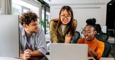 Three coworkers collaborate at a desk in a bright office, looking at a laptop together — one person gestures at the screen while the others listen and engage.