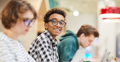 Teenage student wearing glasses smiles toward the camera while seated in a classroom with other students working in the background.