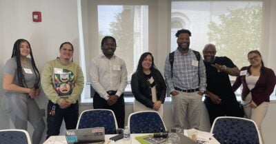 Seven adults stand together behind a conference table during a workshop or meeting. Participants wear name tags and smile toward the camera, with laptops, notebooks, and water bottles visible on the table. Large windows behind the group let in natural light, creating a welcoming professional setting.