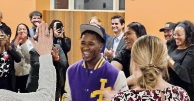 A diverse group of adults gathers indoors, smiling and applauding as a young man in a purple and gold varsity-style jacket and baseball cap shares a joyful moment in the center of the room. Several people laugh, clap, and raise their hands in celebration, creating an energetic and supportive atmosphere.