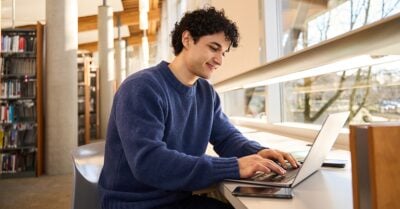 Young man in a blue sweater works on a laptop at a table in a sunlit library.