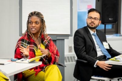 A woman with long braids and a nose ring and a colorful red tartan shawl and yellow pantsuit sits in a classroom, at desks, next to a man in a suit and blue tie.