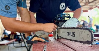 Two people work together to maintain a chainsaw on a workbench, one wearing an AmeriCorps shirt.