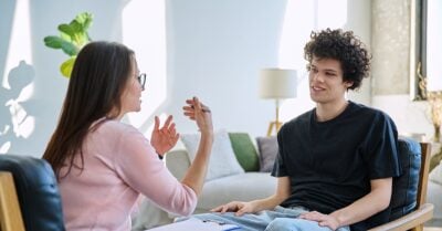 A young man sits in a chair, smiling as he talks with a woman who gestures with her hands.