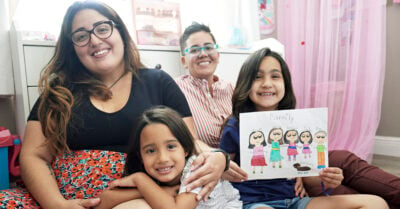A family sits on the floor of a child's room. Two moms sit behind their two daughters. One daughter proudly holds a drawing of her family.