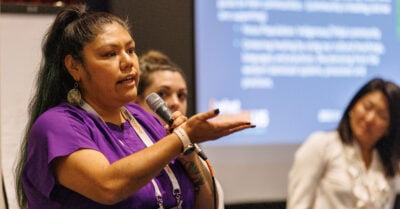 A Latina woman speaks passionately as she presents information seen displayed on a large screen in the background of the right side of the frame. Two other women — one white, one of Asian decent, midground, look on.