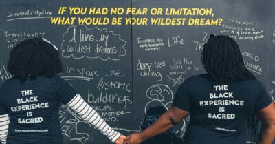 Two Black women stand, holdign hands, in front of a wall filled with handwritten submissions for the Black Thought Project.
