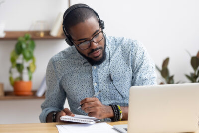 A young black man wearing glasses and a smartly dressed short sleeve dress shirt and headphones sits at a table, pen in hand.