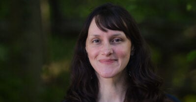 Ellen Roche, a brunette-haired woman, looks at the camera and smiles.