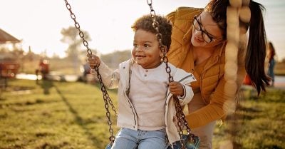 Young Black boy on a swing being supported by an adult female with glasses and a ponytail.