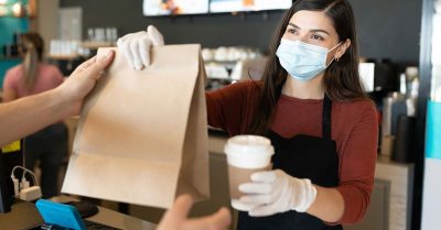 A young female worker at a coffee shop hands a customer their order.
