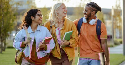 Three college-age students walk outdoors together, smiling and carrying books and backpacks.
