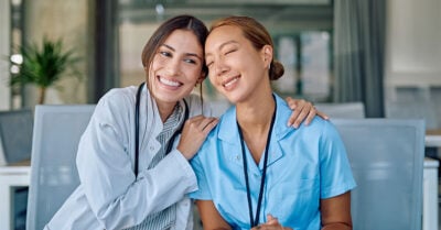 Two young women are wearing blue uniforms. They are seated in an empty lounge and engaged in a side by side embrace. Both women are smiling.