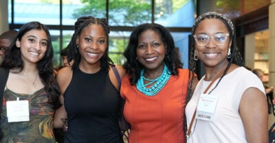 Four women standing together and smiling at a gathering. The woman in the orange dress with a turquoise necklace is centered in the group. Two women stand on her left and one woman on her right.