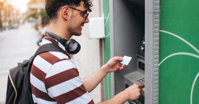 A young man with a black backpack and headphones around his neck stands in front of an ATM. He's smiling and is holding his banking card in front of him, ready to use it at the ATM.