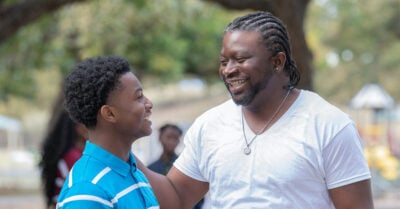 A black father stands outside with his hand on his teenage son's shoulder. Father and son are smiling at one another.