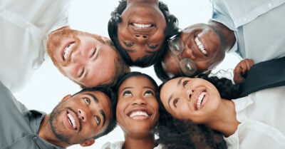 A view of six people taken from below, who are connected in a circle, interlocked arm and arm, and smiling.