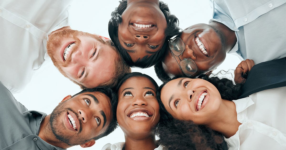 A view of six people taken from below, who are connected in a circle, interlocked arm and arm, and smiling.