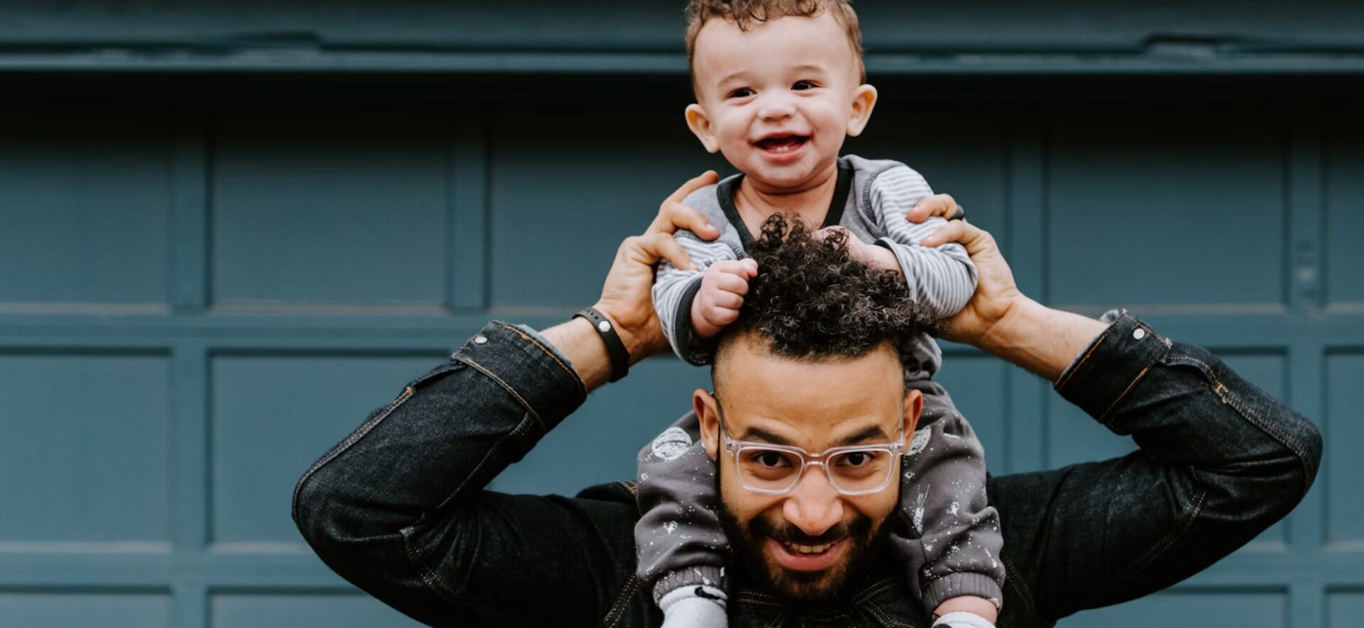 Hispanic male with denim jacket and glasses holds young child on his shoulders while the infant plays with dad's curly hair.
