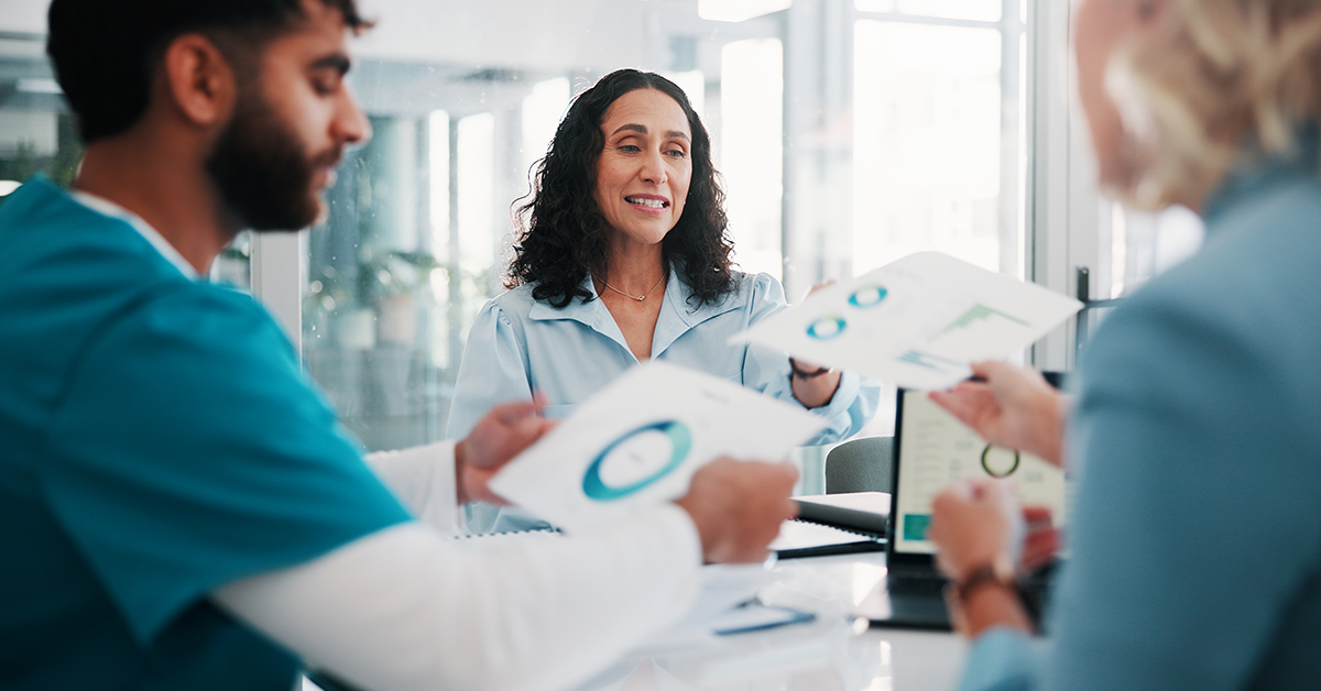 A woman sits in an office setting between two other people, and they are passing papers to one another that contain charts and graphs.