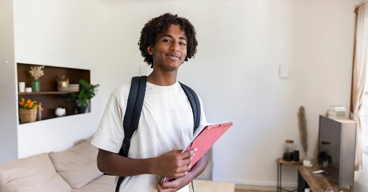A young person wearing a backpack holds a pink notebook and smiles at the camera.