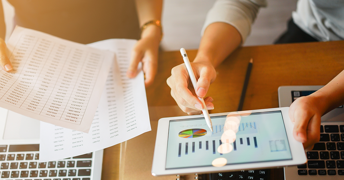 A close up view of two people pouring over data in the form of hardcopy spreadsheets and an iPad with charts on it. Laptops sit on the table beneath them.